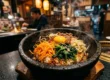 Close-up detalhado de um Bibimbap em tigela de pedra aquecida, destacando as cores vibrantes dos vegetais e a gema de ovo central em ambiente de restaurante japonês refinado