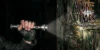 Close de personagem em jogo de terror segurando uma lanterna acesa em um corredor escuro, com o feixe de luz destacando detalhes de uma parede rústica de madeira e uma moldura de quadro antiga no canto direito.